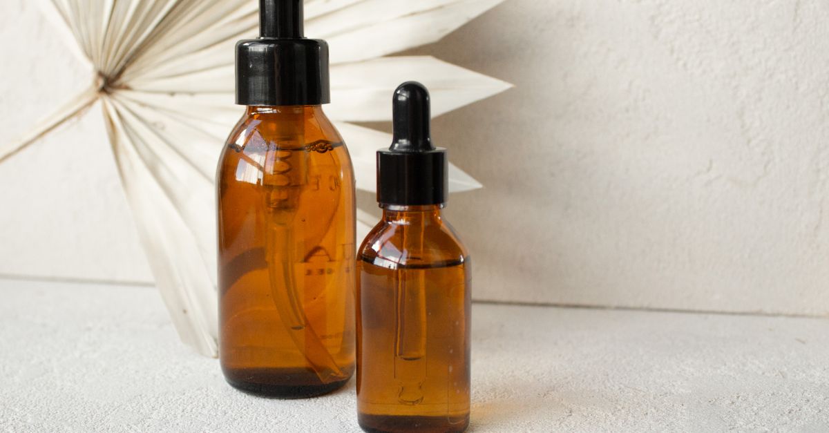 Two amber glass dropper bottles stand side by side on a pale textured surface, a folded paper fan behind them.