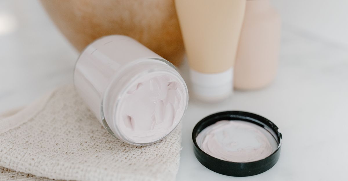 A glass jar of thick pale cream lies tipped on its side on a woven cloth, its black lid resting open beside it.