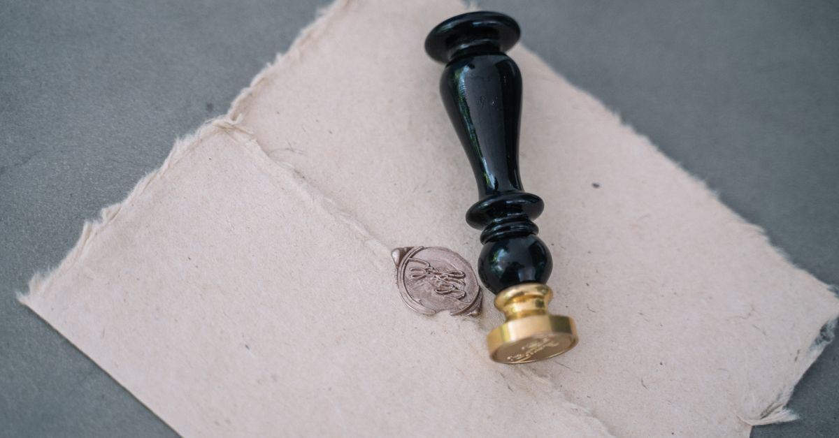 A black and brass wax seal stamp rests on handmade paper beside the grey impression it just left.