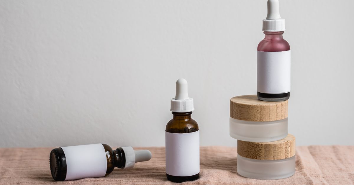 Three cosmetic bottles with blank white labels: one lying flat on wood, one upright, one standing on a stack of frosted jars with bamboo lids.