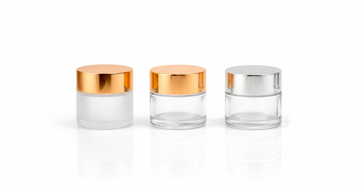 Three small glass cream jars, two with gold lids and one with a silver lid, stand in a row on a white surface.