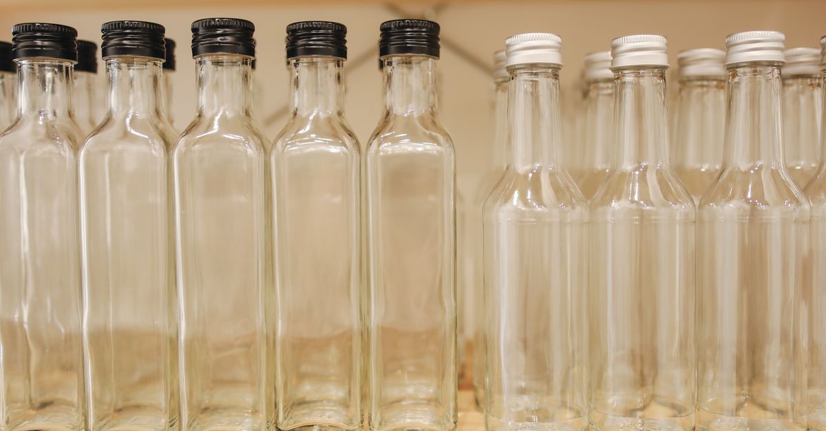 Two rows of empty clear glass bottles stand lined up on a wooden shelf, dark caps on one row and white caps on the other.