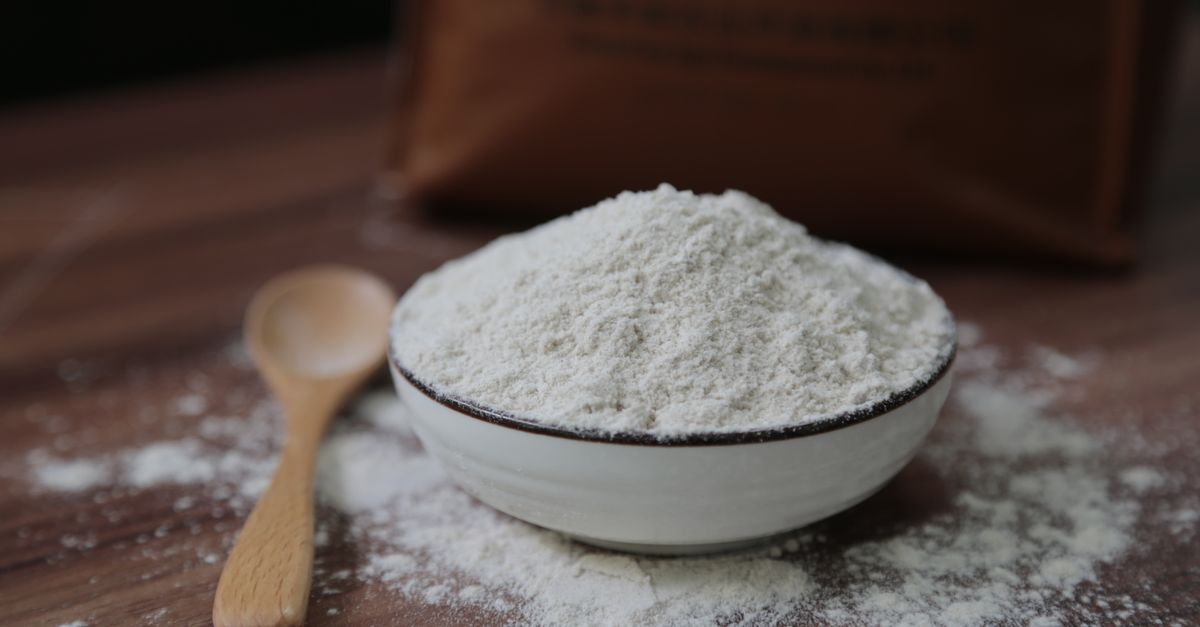 A white bowl heaped with fine pale powder sits on dark wood beside a small wooden spoon, powder dusted across the table.