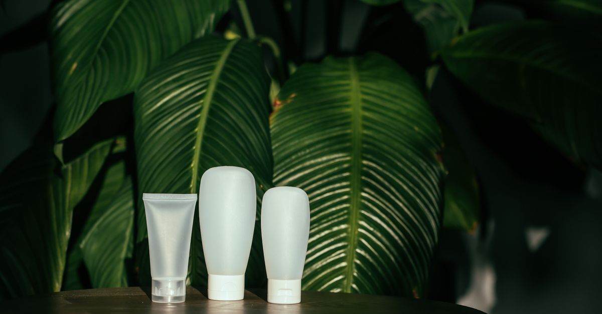 Three white frosted tubes and bottles stand in a row on a wooden table in front of dark green leaves.