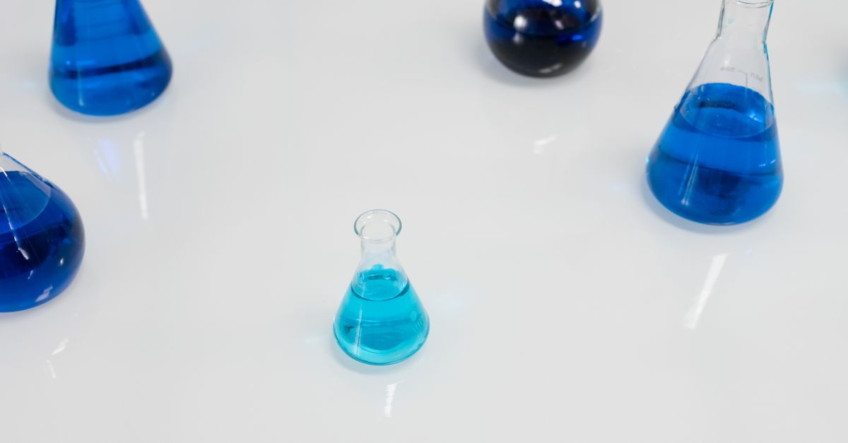 Four laboratory flasks holding blue liquid of different depths stand on a white surface.