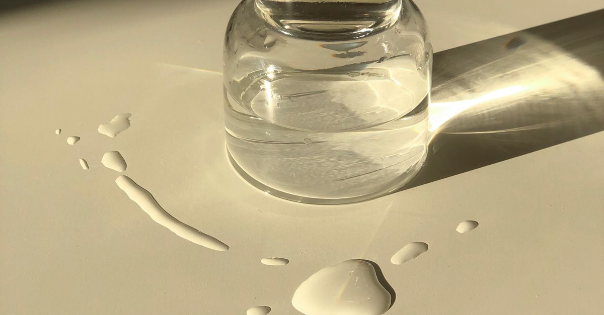 A small round glass jar of clear liquid casts a long shadow on a warm beige surface, with drops spilled beside it.