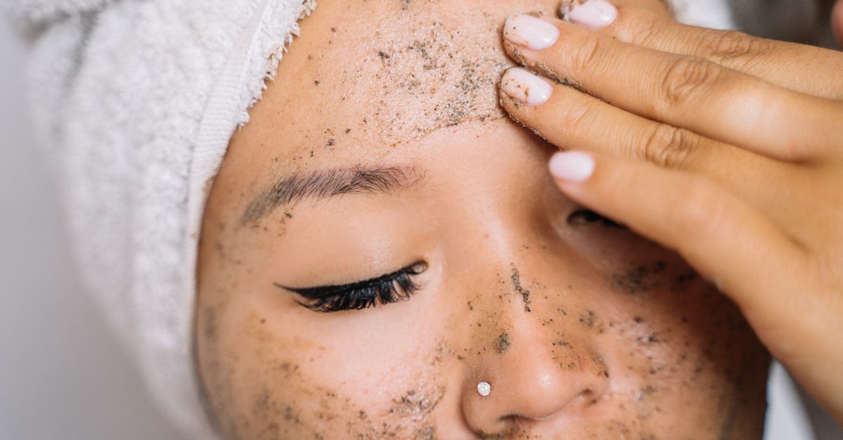 A person with a towel wrapped around their hair rubs a white scrub across one cheek.