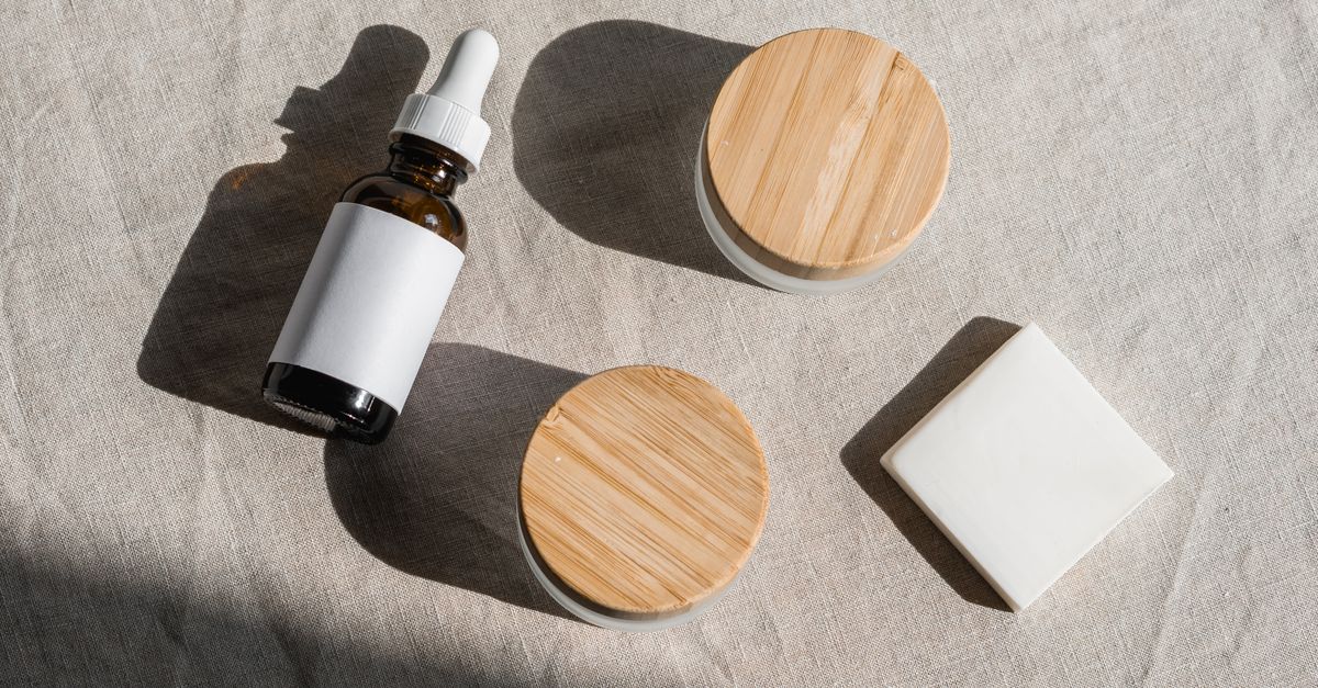 A dropper bottle, two jars with bamboo lids and a square bar lie on linen cloth in hard sunlight.