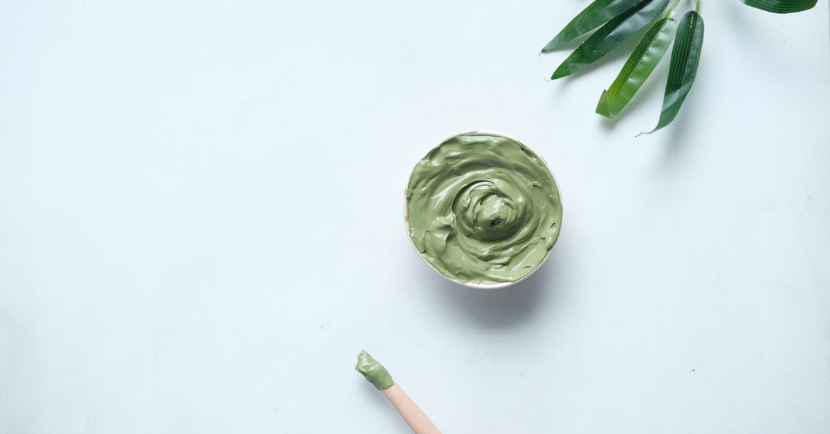 A small dish of swirled green clay sits on a pale blue surface with a wooden spatula and green leaves beside it.