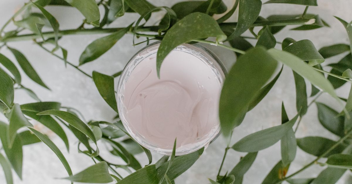 A round glass jar of pale pink cream with thin green leafy branches drooping over it.
