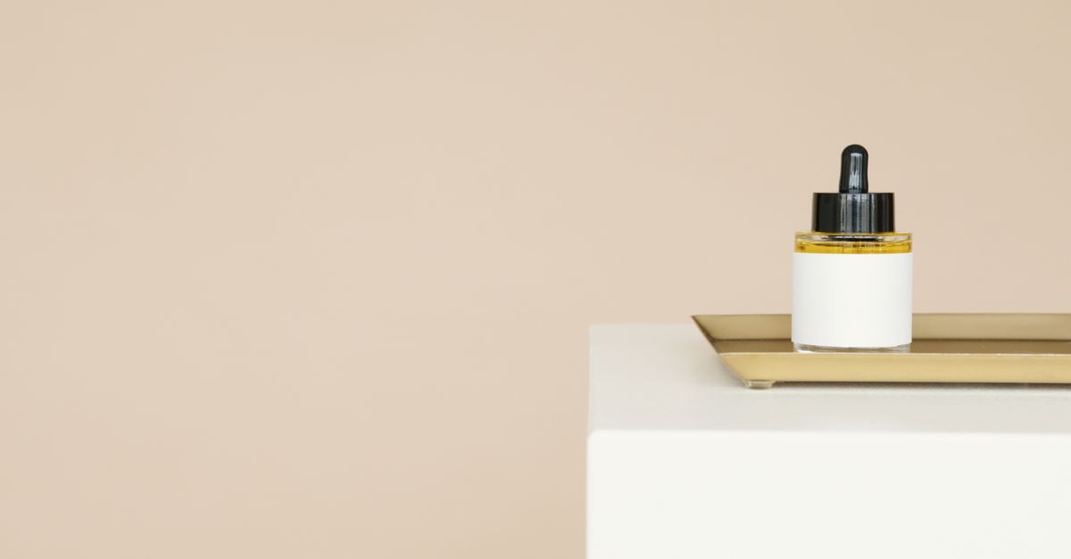 A small dropper bottle sits on a gold tray on a white plinth against a warm beige background.