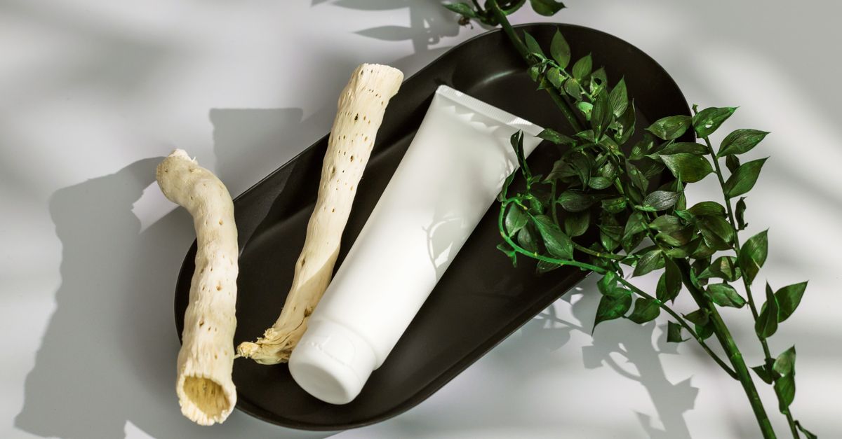 A white tube lies on a dark oval plate with a dried root and green sprigs around it.