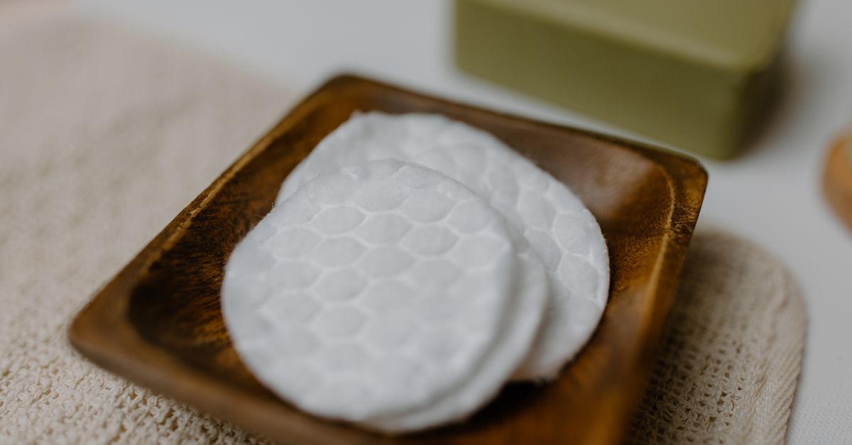 A stack of round embossed cotton pads sits on a dark wooden dish, a green bar behind it.