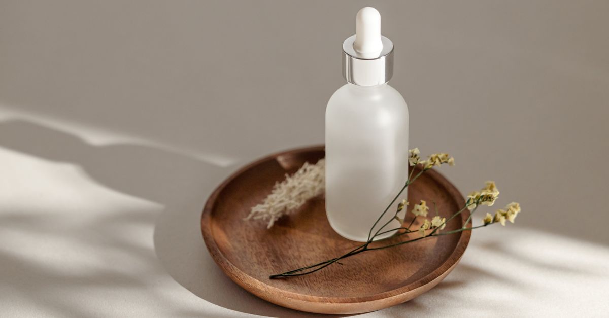 A frosted white dropper bottle stands on a wooden plate with dried flowers beside it.