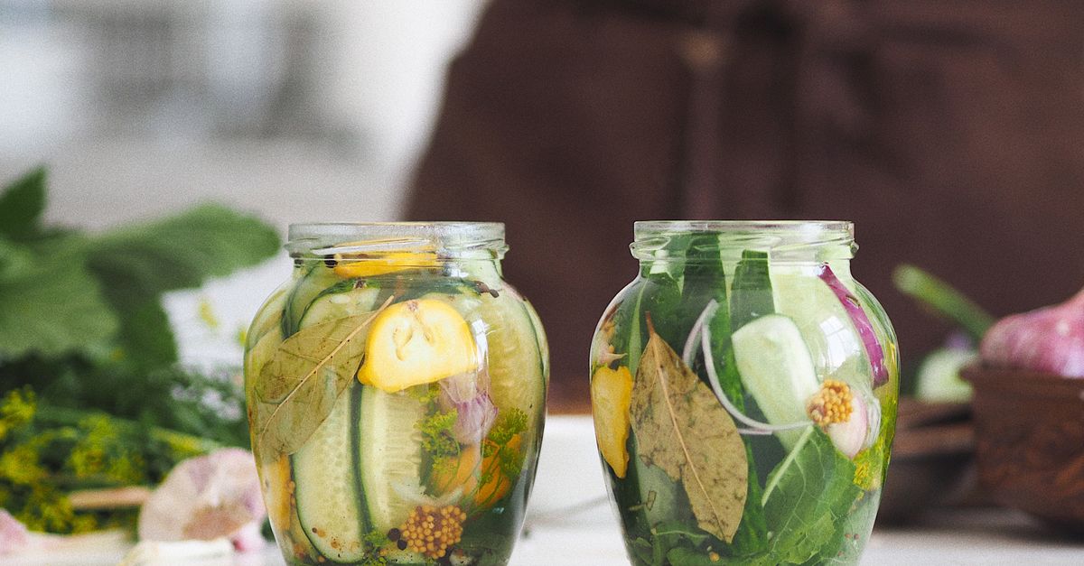 Two glass jars of pickled cucumber and lemon with herbs stand on a kitchen counter.