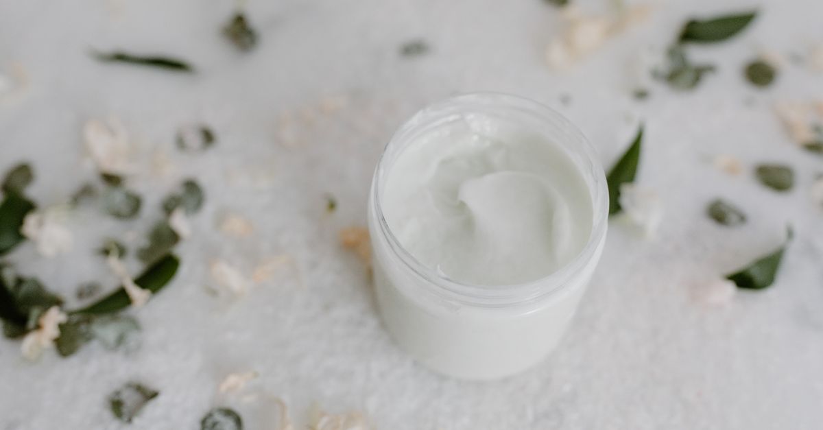 An open white jar of thick cream sits on a pale surface scattered with small leaves and petals.