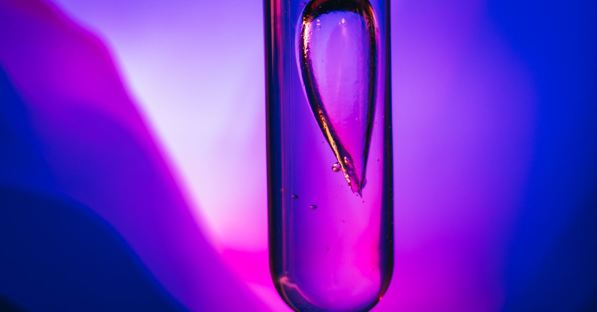 A glass test tube holds a single teardrop of liquid, lit in violet and magenta.