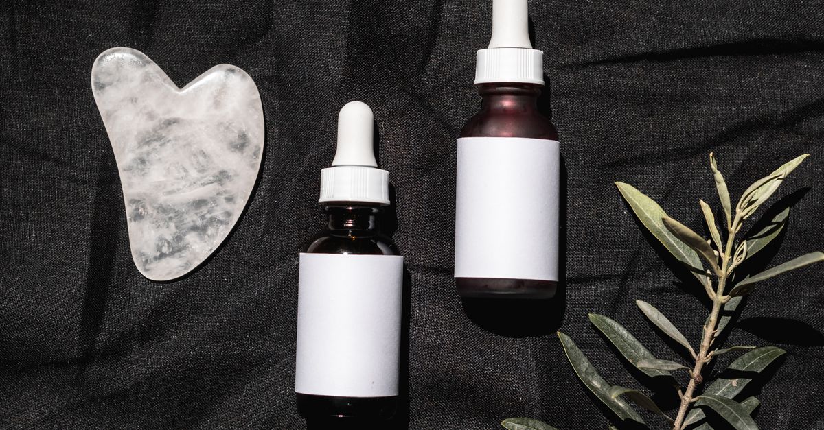Two dropper bottles with blank labels lie on black cloth with a heart shaped stone tool and an olive sprig.