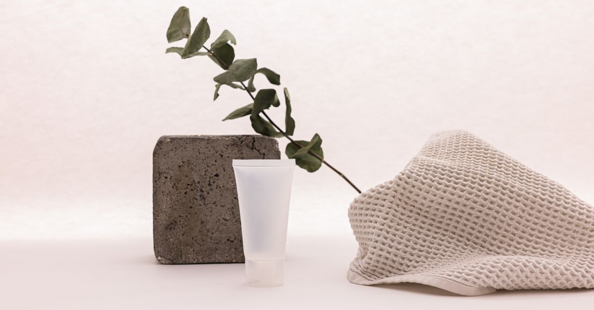 A small frosted tube stands between a grey stone block and folded waffle cloth, a eucalyptus sprig behind, on a pink ground.