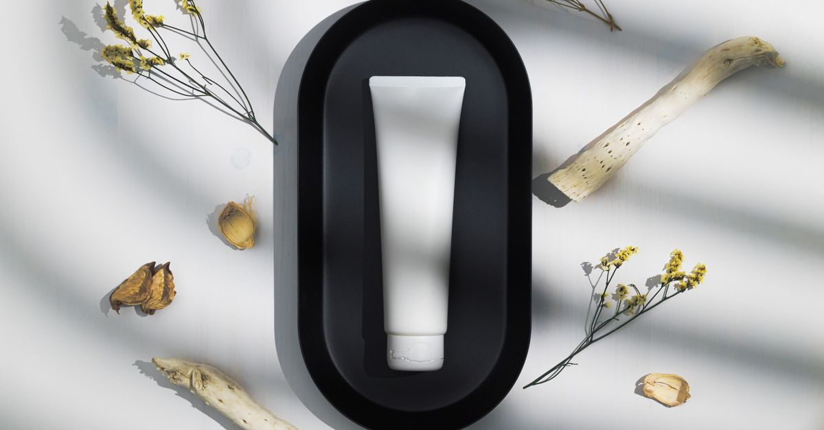 A white tube sits inside a black oval frame on a white surface, dried flowers and roots scattered around it.