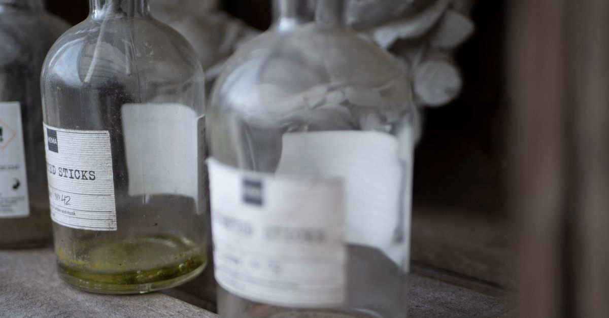 Two large glass apothecary bottles with paper labels stand on a bench, one holding olive coloured liquid.