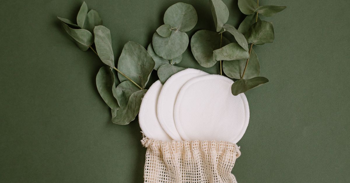 Three round cotton pads spill from a mesh pouch with eucalyptus branches above, on a deep green ground.