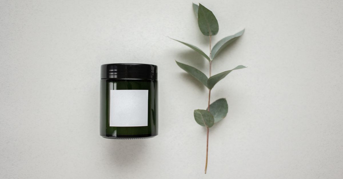 A dark glass jar with a blank white label sits beside a eucalyptus sprig on a pale grey surface.