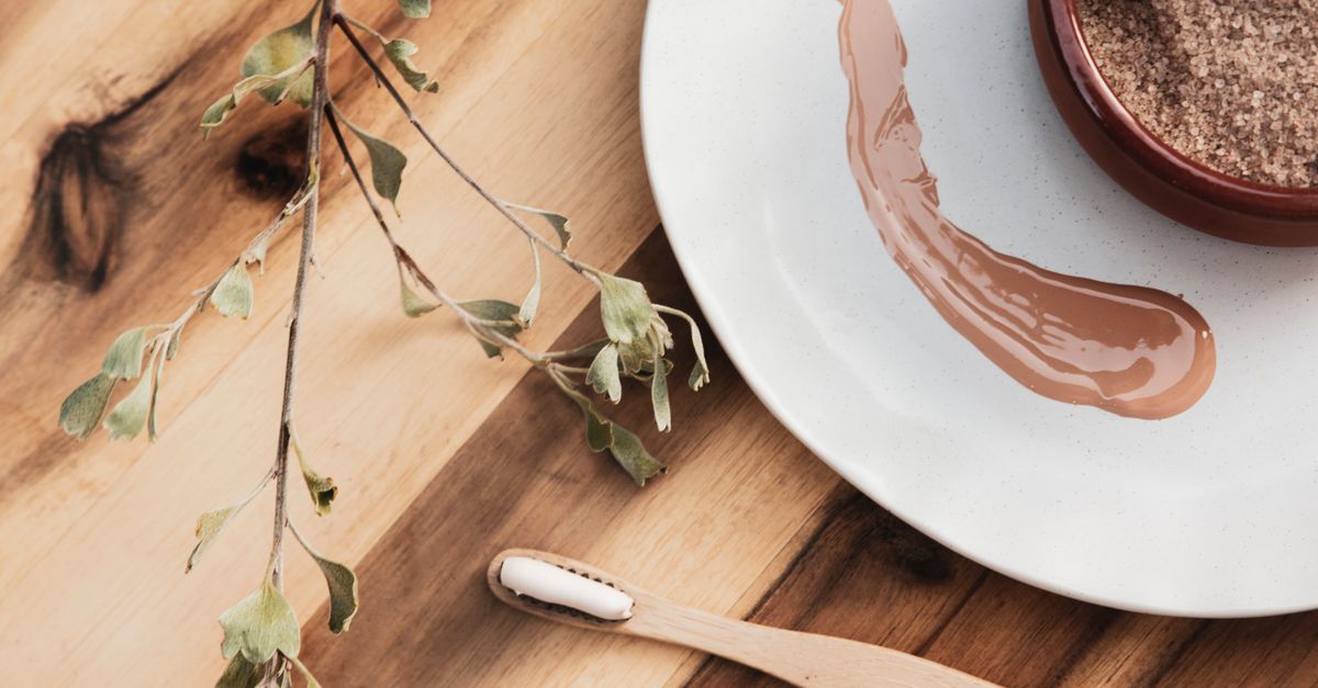 A plate carries a swipe of clay coloured mask beside a wooden spatula and a bowl of dry powder on a wooden board.