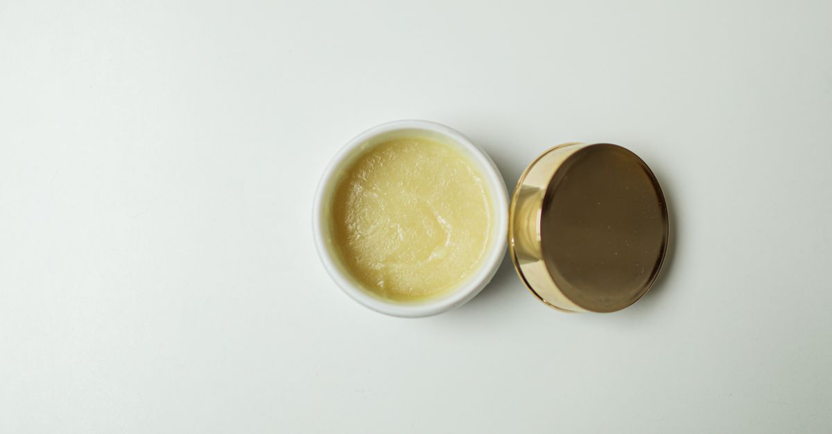 An open jar of pale yellow balm sits on a pale green surface with its gold lid beside it.