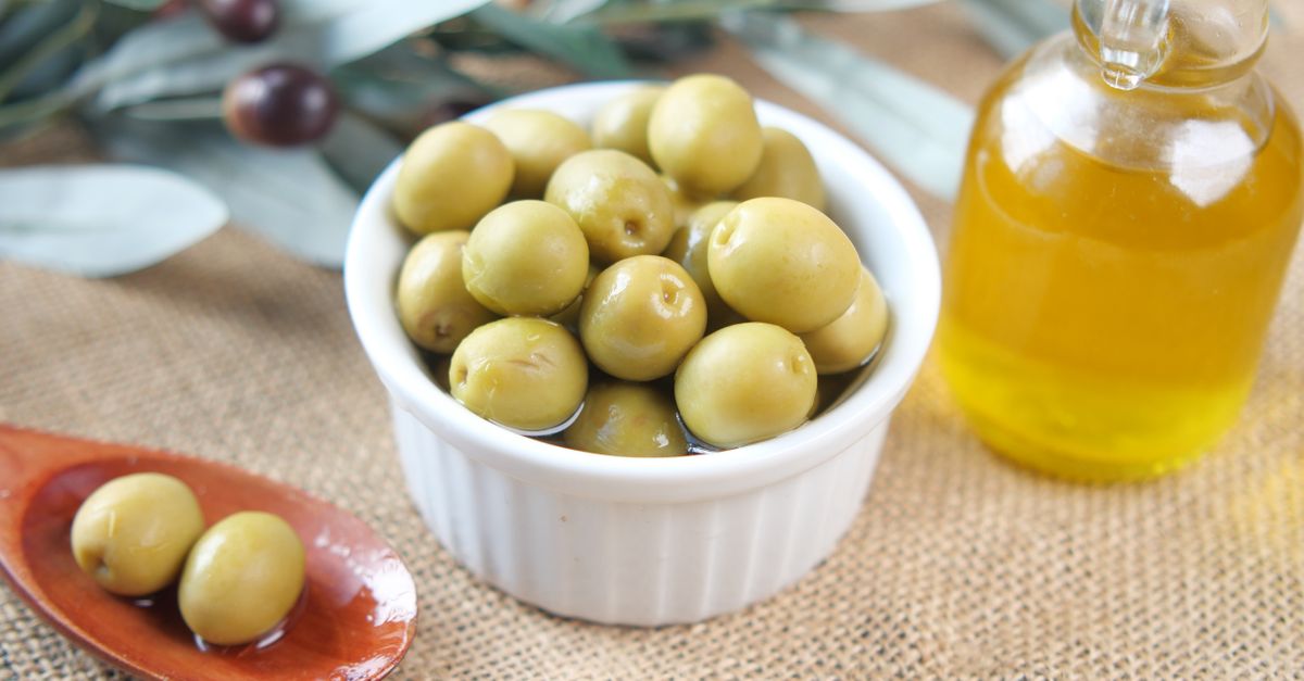 A bowl of green olives stands beside a glass bottle of golden oil on burlap.