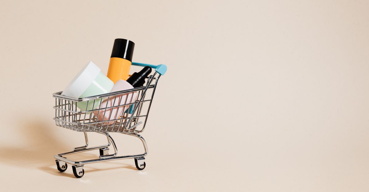 A miniature shopping trolley is loaded with cosmetics on a plain beige ground.